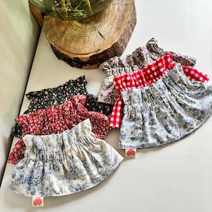 May include: A collection of ruffled fabric skirts in various floral and patterned designs. The skirts feature colors like red, black, blue, and white. Each skirt has a small heart-shaped tag attached.