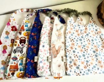 May include: Six fabric baby bibs with elastic neckbands, featuring various patterns including cartoon characters, cars, blue floral motifs, and small pink and blue flowers on white, blue, and cream backgrounds, arranged in a row on a surface.