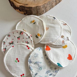 May include: A selection of round, white fabric pads with diverse printed designs. Some pads feature floral patterns, cherries, lemons, birds, and hearts. The pads have a white, textured border and are arranged on a white surface.