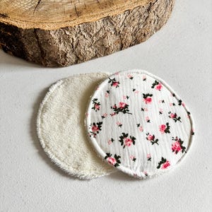 May include: Two reusable facial rounds. One is white terry cloth, the other is white with a pink floral pattern. The rounds are circular and sit on a white surface, with a wooden slice in the background.