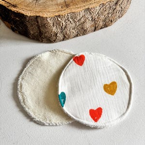 May include: Two reusable facial rounds. One is cream-coloured, the other is white with colourful heart designs. The hearts are red, teal, and gold. The rounds are made of a soft, textured material.