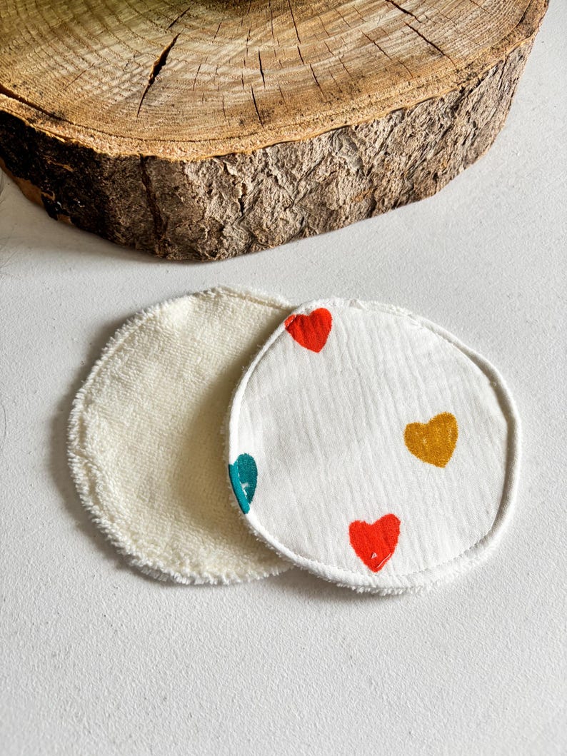 May include: Two reusable facial rounds. One is cream-coloured, the other is white with colourful heart designs. The hearts are red, teal, and gold. The rounds are made of a soft, textured material.