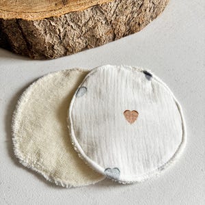 May include: Two circular reusable facial pads. One is a soft, cream-coloured terry cloth. The other is white with a stitched gold heart and small grey hearts. These pads are designed for skincare.