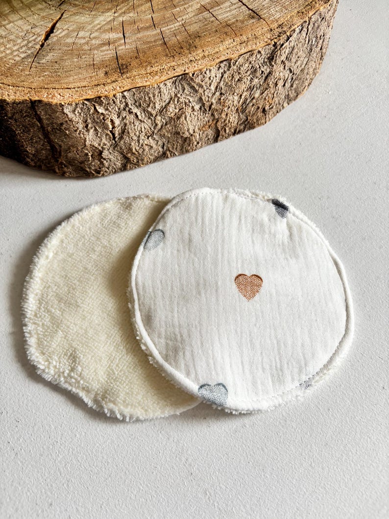 May include: Two circular reusable facial pads. One is a soft, cream-coloured terry cloth. The other is white with a stitched gold heart and small grey hearts. These pads are designed for skincare.