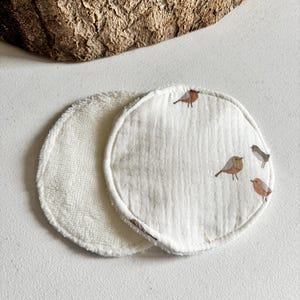 May include: Two circular reusable facial cleansing pads. One is white terry cloth, the other is white muslin with a bird pattern in brown and grey. These pads are designed for gentle skincare and makeup removal.