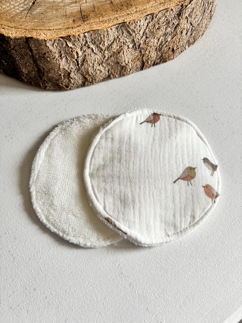 May include: Two circular reusable facial cleansing pads. One is white terry cloth, the other is white muslin with a bird pattern in brown and grey. These pads are designed for gentle skincare and makeup removal.