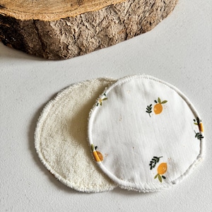 May include: Two reusable facial rounds. One is a soft, cream-coloured terry cloth. The other is white with a pattern of orange lemons and green leaves. The rounds are circular and have a stitched edge.