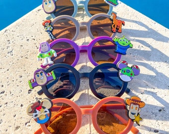 May include: A stack of six colorful sunglasses with cartoon character charms. The sunglasses are in various colors, including purple, blue, black, and orange. The charms include characters from the Toy Story franchise, such as Woody, Buzz Lightyear, and Rex.