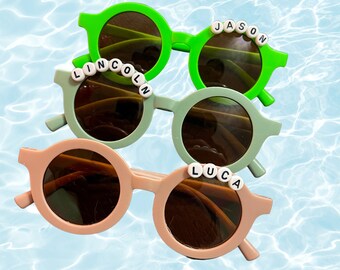 May include: Four pairs of round sunglasses in green, sage green, and peach. Each pair has a name spelled out in white letter beads across the top of the frame: "JASON", "LINCOLN", and "LUCA". The background is a light blue water pattern.