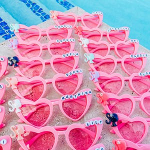May include: Pink heart-shaped sunglasses with a Barbie doll charm and personalized names in white letters. The sunglasses are arranged in a pile on a light brown surface.