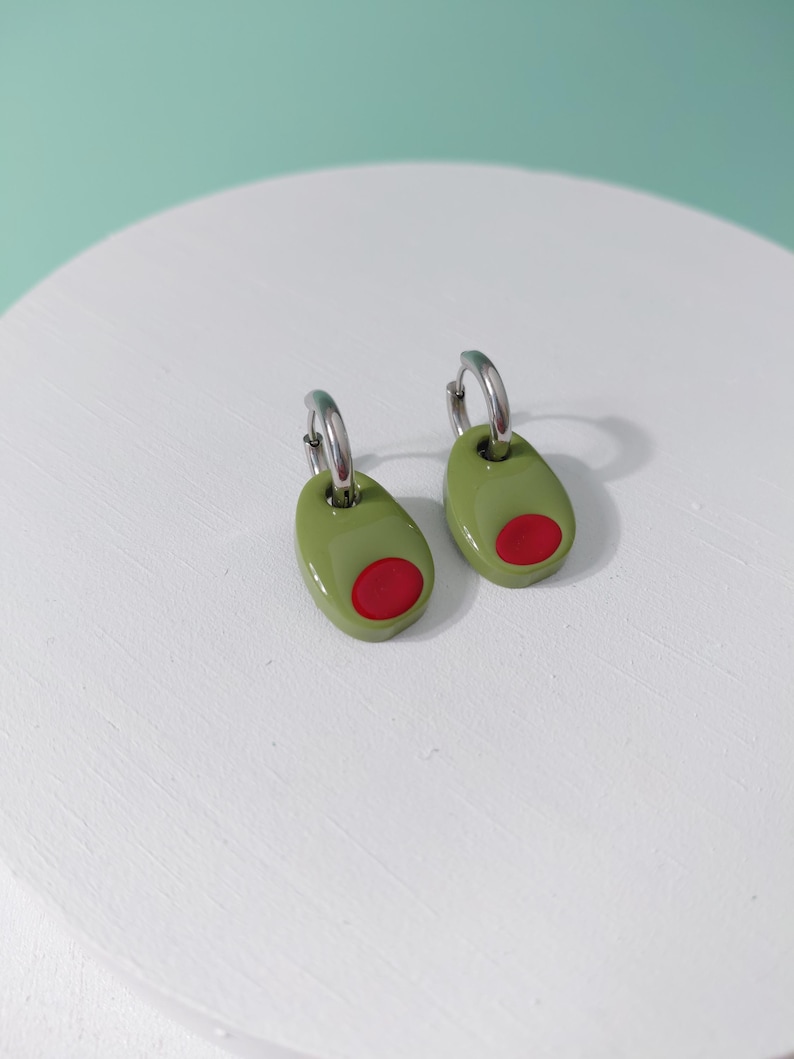 K&ouml;nnte beinhalten: Ein Paar olivenf&ouml;rmige Ohrringe. Jeder Ohrring ist gr&uuml;n mit einem roten Zentrum, das einer Olive &auml;hnelt. Die Ohrringe haben silberfarbene Reifen zum Befestigen.