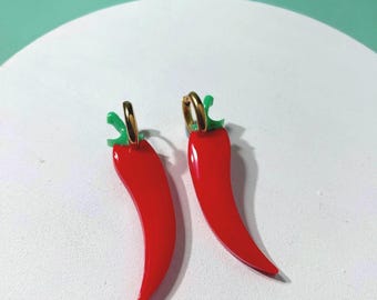 May include: A pair of hoop earrings featuring bright red chilli pepper charms with small green stems. The earrings are constructed with gold-toned metal hoops that connect to the red pepper pendants, creating a vibrant and distinct accessory set.