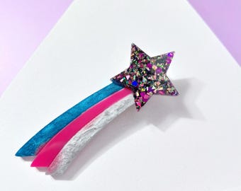 May include: A star-shaped brooch with a glittery, multi-coloured design. The brooch has a blue, pink, and white trail. The brooch is set against a white triangular background with a purple backdrop.
