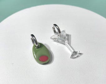 May include: A pair of mismatched hoop earrings featuring a silver-tone metal hoop attached to a green olive charm with a red centre, and a second silver-tone hoop attached to a clear, transparent martini glass-shaped charm.