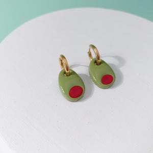 K&ouml;nnte beinhalten: Ein Paar Ohrringe in Olivenform. Jeder Ohrring hat einen goldenen Reifen und ein gr&uuml;nes Oval mit einem roten Kreis, der einer Olive &auml;hnelt. Die Ohrringe werden auf einer wei&szlig;en Oberfl&auml;che vor einem hellgr&uuml;nen Hintergrund pr&auml;sentiert.
