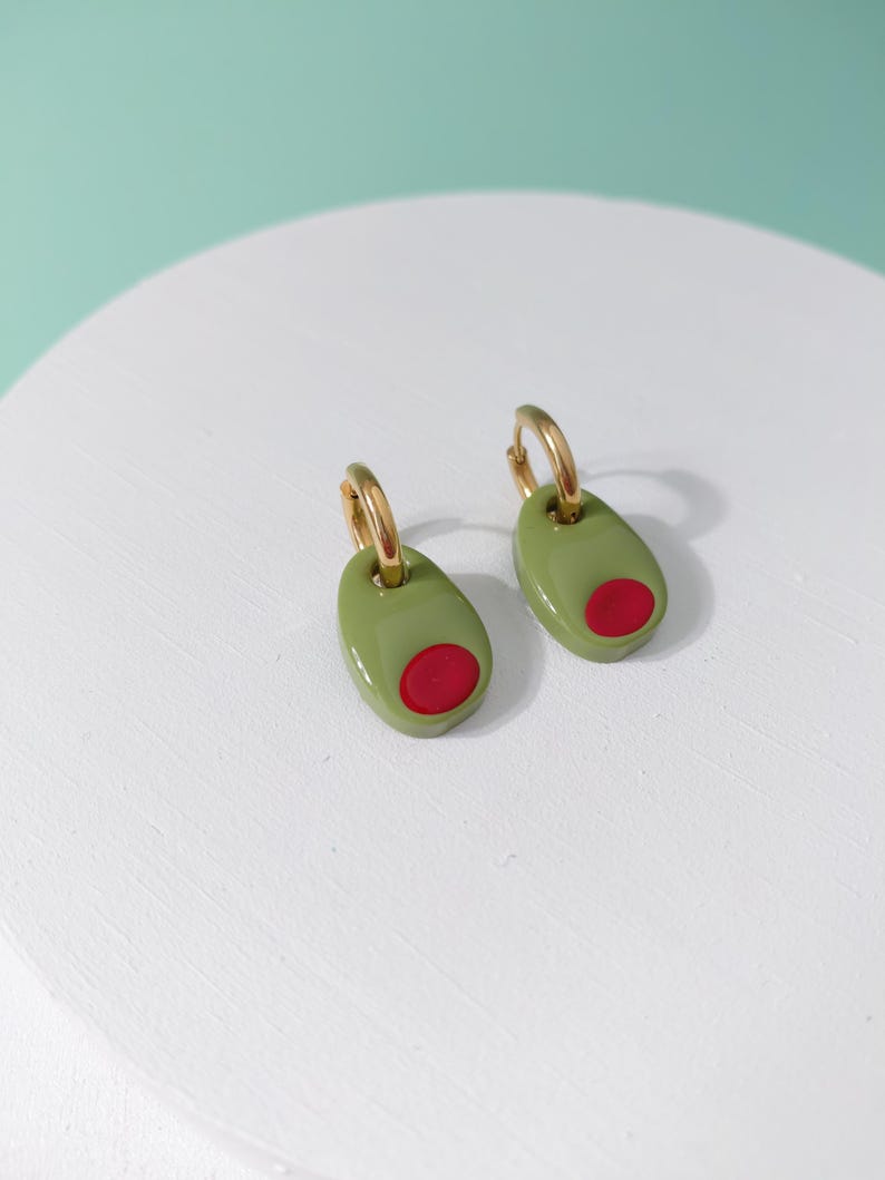 K&ouml;nnte beinhalten: Ein Paar Ohrringe in Olivenform. Jeder Ohrring hat einen goldenen Reifen und ein gr&uuml;nes Oval mit einem roten Kreis, der einer Olive &auml;hnelt. Die Ohrringe werden auf einer wei&szlig;en Oberfl&auml;che vor einem hellgr&uuml;nen Hintergrund pr&auml;sentiert.
