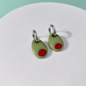K&ouml;nnte beinhalten: Ein Paar olivenf&ouml;rmige Ohrringe. Jeder Ohrring hat ein gr&uuml;nes Oval mit einem roten Kreis, der einer Olive nachempfunden ist. Die Ohrringe sind an silberfarbenen Creolen befestigt. Die Ohrringe werden auf einer wei&szlig;en Oberfl&auml;che pr&auml;sentiert.