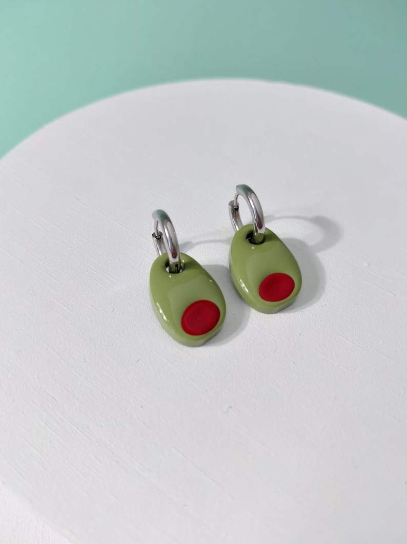 K&ouml;nnte beinhalten: Ein Paar olivenf&ouml;rmige Ohrringe. Jeder Ohrring hat ein gr&uuml;nes Oval mit einem roten Kreis, der einer Olive nachempfunden ist. Die Ohrringe sind an silberfarbenen Creolen befestigt. Die Ohrringe werden auf einer wei&szlig;en Oberfl&auml;che pr&auml;sentiert.