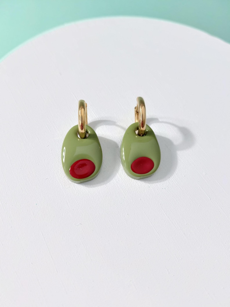 K&ouml;nnte beinhalten: Olivenf&ouml;rmige Ohrringe. Jeder Ohrring hat ein gr&uuml;nes Oval mit einem roten Kern, das einer Olive &auml;hnelt. Die Ohrringe h&auml;ngen an goldfarbenen Creolen und sind ein verspieltes und einzigartiges Accessoire.