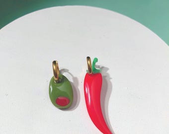 May include: A pair of mismatched earrings featuring a gold-tone hoop with a green olive charm containing a red pimento centre, and a gold-tone hoop with a bright red chilli pepper charm featuring a small green stem at the top.