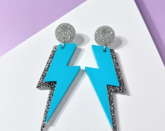May include: Turquoise lightning bolt earrings with silver glitter detailing. Each earring features a silver glitter disc at the top. The earrings are displayed against a white and purple backdrop.