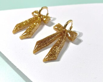 May include: A pair of gold glitter bow earrings featuring small, polished gold-tone hoop closures. The bows are crafted with a dense, sparkling gold finish and are suspended from the circular hoops, creating a dangling effect for the ears.