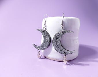 May include: Silver glitter crescent moon earrings with star charms. The earrings are displayed on a white ceramic cylinder against a purple background. The earrings have silver hooks. The earrings are a celestial themed accessory.