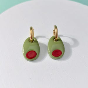 K&ouml;nnte beinhalten: Ein Paar olivf&ouml;rmige Ohrringe. Jeder Ohrring hat einen goldfarbenen B&uuml;gel und einen gr&uuml;nen ovalen Anh&auml;nger mit roter Mitte, der einer Olive &auml;hnelt. Die Ohrringe werden auf einer wei&szlig;en Oberfl&auml;che pr&auml;sentiert.
