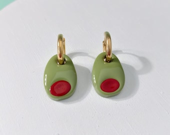 May include: A pair of olive-shaped earrings. Each earring has a gold-coloured hoop and a green oval pendant with a red centre, resembling an olive. The earrings are displayed on a white surface.