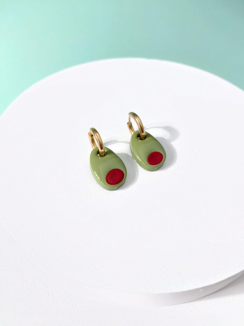 K&ouml;nnte beinhalten: Ein Paar olivf&ouml;rmige Ohrringe. Jeder Ohrring hat ein gr&uuml;nes Oval mit einem roten Punkt, das einer Olive &auml;hnelt. Die Ohrringe haben goldfarbene Creolenverschl&uuml;sse und sind ein verspieltes und einzigartiges Accessoire.