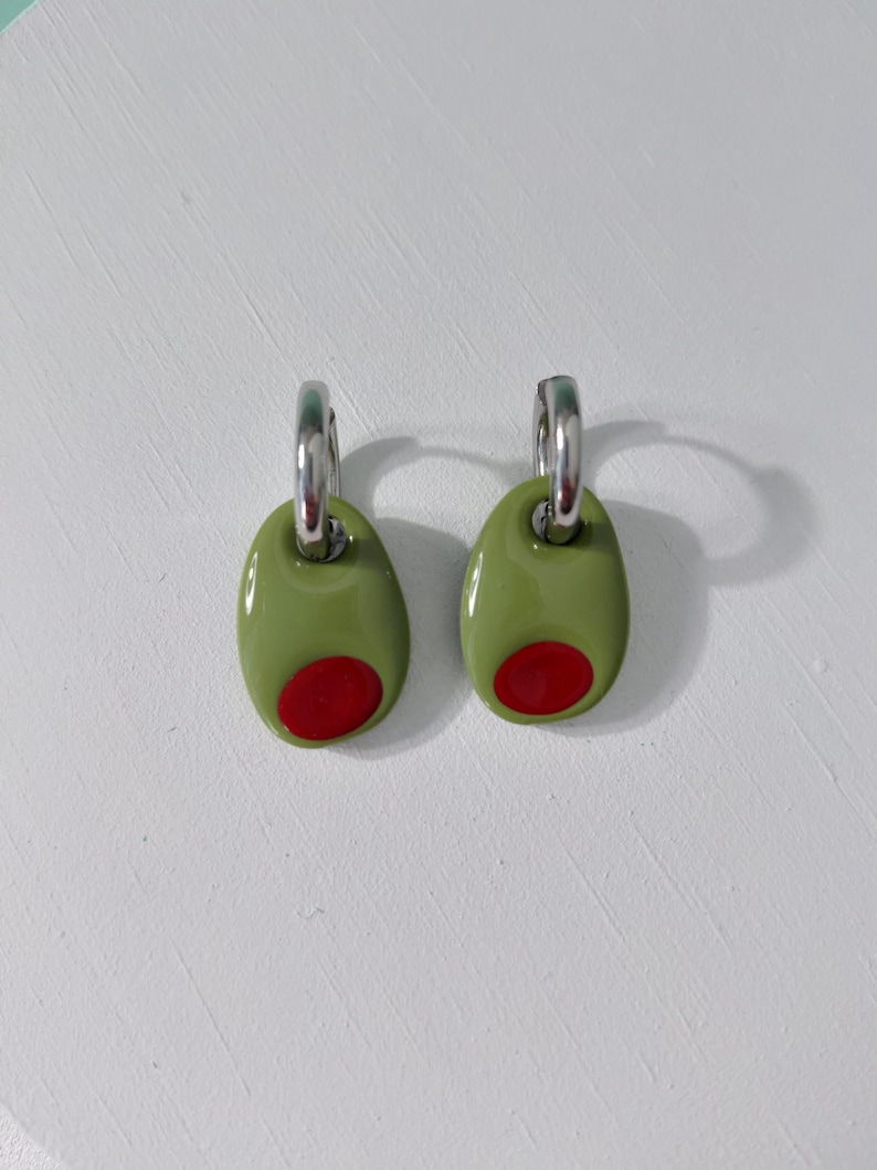 K&ouml;nnte beinhalten: Ein Paar olivf&ouml;rmige Ohrringe. Jeder Ohrring hat ein gr&uuml;nes Oval mit einem roten Punkt, das einer Olive &auml;hnelt. Die Ohrringe haben silberfarbene Schlaufen zur Befestigung.