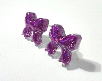 May include: A pair of stud earrings shaped like bows, crafted from a translucent purple resin material embedded with fine, reflective glitter particles that catch the light, featuring a standard post backing for secure attachment.