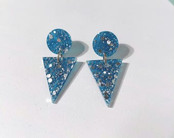 May include: A pair of blue resin earrings with silver glitter. Each earring has a circular top and a triangular pendant. The earrings are displayed on a white surface with a light blue background.