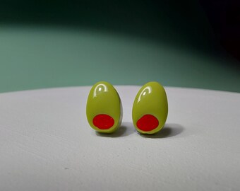 May include: A pair of olive-shaped stud earrings. Each earring is a glossy, light green oval with a bright red centre. The earrings are set against a white surface with a soft green background.