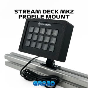May include: A black Stream Deck MK2 profile mount with a silver metal track. The Stream Deck has 15 buttons with clear labels. The text "STREAM DECK" is printed on the top of the device.