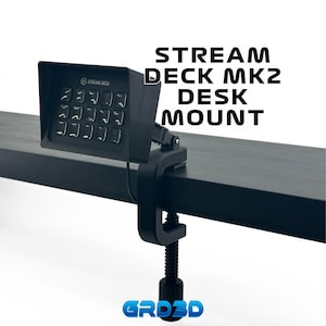 May include: Black Stream Deck MK2 desk mount with a clamp. The mount is attached to a black surface. The Stream Deck MK2 is a device with a grid of 15 buttons.
