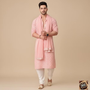 May include: A light pink kurta pajama set with a matching scarf. The kurta features intricate embroidery and is paired with white pants. The scarf has a gold border. The outfit is worn with matching shoes.