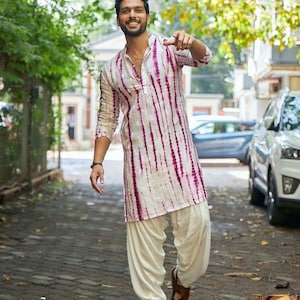 Puede incluir: Un hombre con una kurta blanca con rayas verticales teñidas con tinte morado, combinada con pantalones salwar color crema y sandalias marrones. El atuendo se usa al aire libre en un camino de ladrillos, con árboles y coches al fondo.
