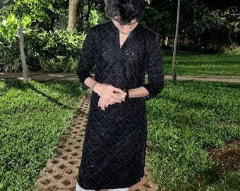 May include: A man wearing a black long-sleeved shirt with a sparkly pattern and white pants. He is standing on a brick pathway in a garden setting.