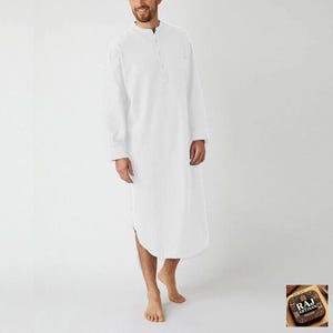May include: A long-sleeved white tunic featuring a mandarin collar, a partial button-down placket, and a single chest pocket. The garment reaches below the knees and has a curved hemline, providing a clean, solid-coloured appearance.
