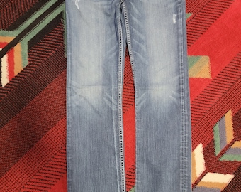 May include: Light blue denim jeans with a faded wash and distressed details. The jeans have a classic five-pocket design, a zip fly, and white stitching. The jeans are laid flat on a patterned red and black rug.