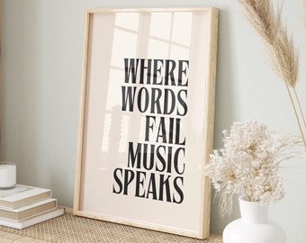 May include: A framed print with the text "Where words fail music speaks" in black lettering on a light pink background. The print is displayed on a light wood shelf with a white vase and dried flowers.