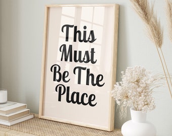 May include: A framed print with the text "This Must Be The Place" in black lettering on a light pink background. The print is hanging on a wall above a wooden bench with woven rattan seats. There are two woven baskets and a white vase with dried flowers on the bench.