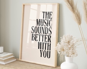 May include: A framed print with the text "THE MUSIC SOUNDS BETTER WITH YOU" in black lettering on a light pink background. The print is displayed on a light wood shelf with a woven rattan surface. There are two woven baskets and a white vase with dried flowers on the shelf.