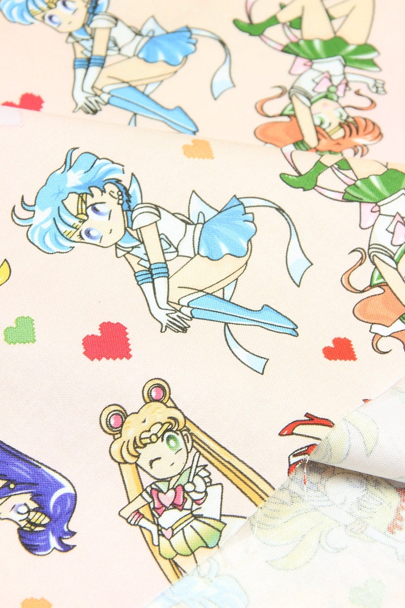 May include: Pink fabric with a repeating pattern of anime characters in various poses. The characters are wearing sailor outfits with different colored accents. The fabric is decorated with small hearts.