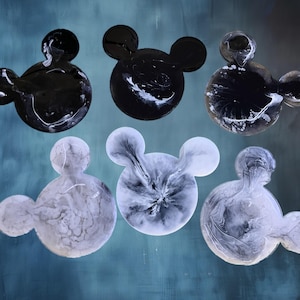 May include: Six Mickey Mouse-shaped decorative pieces. Three are black with white marbling, and three are white with gray marbling. The pieces have a glossy finish and are arranged on a teal-colored background.