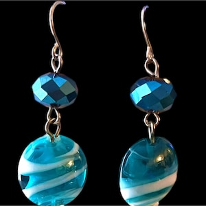 May include: A pair of dangle earrings with a dark background. Each earring features a faceted, teal-colored bead above a larger, oval bead with white swirls. The earrings have silver-colored hooks and connecting links.