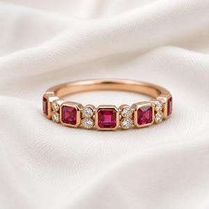 May include: A rose gold band ring featuring a repeating pattern of square-cut red rubies and small round white diamonds. The gemstones are set in a bezel style along the top half of the metal band, creating a symmetrical and alternating design.