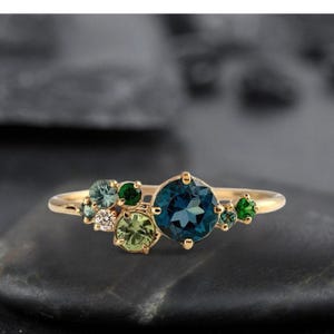 May include: A gold ring featuring a cluster of gemstones. The ring has a large, round, deep blue center stone, surrounded by smaller green and clear stones. The band is a thin, polished gold.
