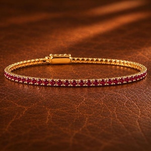 May include: A gold tennis bracelet featuring a continuous row of small, round, vibrant red rubies set in a polished gold metal frame. The bracelet includes a rectangular gold clasp mechanism at the top, showcasing a sleek and uniform design.